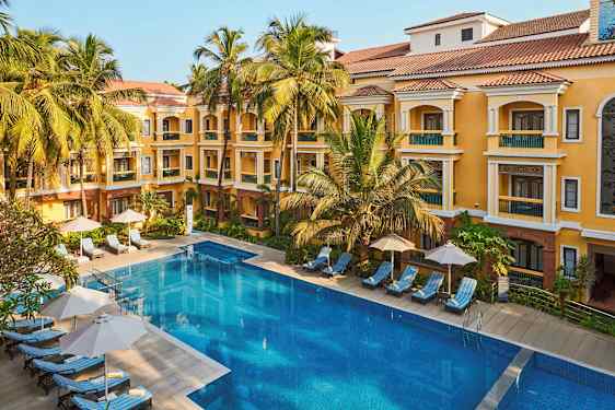 En bild av en hotellbyggnad med gul fasad och röda tak, omgiven av palmer. Framför byggnaden finns en stor pool och solstolar med parasoller. (AI-genererad text)