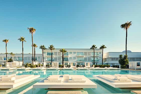 En modern byggnad med balkonger, omgiven av palmer och en swimmingpool med solstolar. (AI-genererad text)