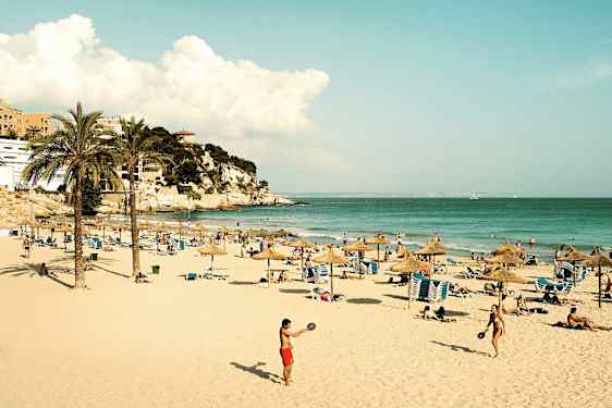 En sandstrand med parasoller, solstolar och människor som njuter av solen och havet. Längre bort syns en klippa med byggnader. (AI-genererad text)