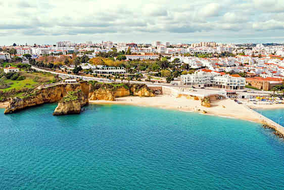 Ljus sandstrand vid havet med en stad i bakgrunden och klippor intill stranden. (AI-genererad text)