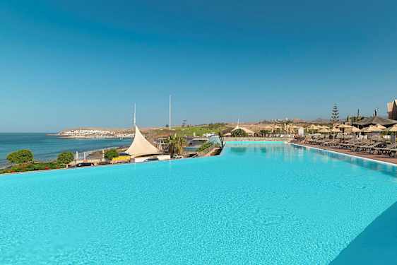 En stor utomhuspool med solstolar och parasoll bredvid, med utsikt över havet och en kuststad. (AI-genererad text)