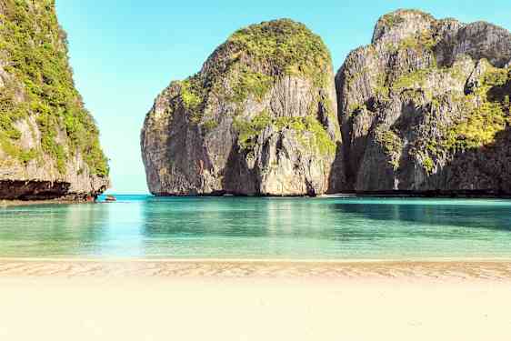 En sandstrand som vetter mot lugnt blått vatten, omgiven av höga klippor täckta av vegetation. (AI-genererad text)