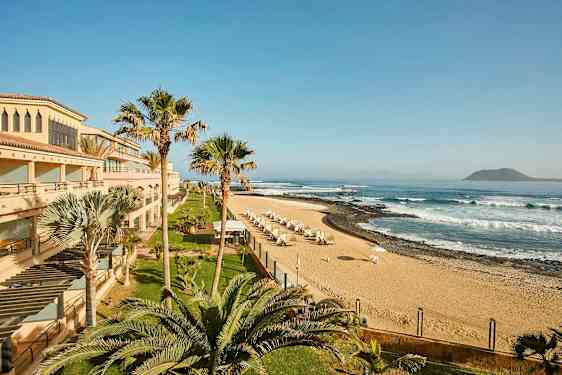 En strand med sand, parasoller och vågor, omgiven av byggnader och palmer. (AI-genererad text)