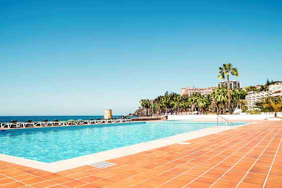 En swimmingpool med omgivande solsängar och vy över havet och bebyggelse med palmer. (AI-genererad text)