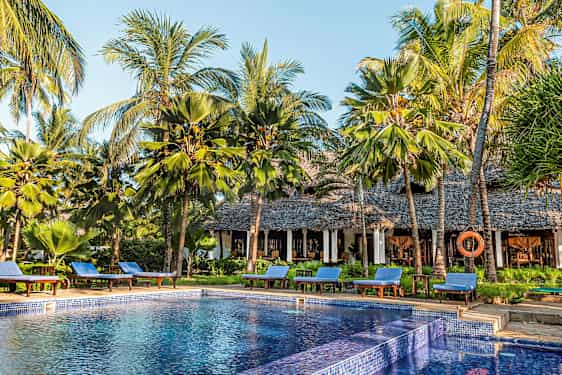 En pool omgiven av solstolar och palmer vid en byggnad med stråtak. (AI-genererad text)