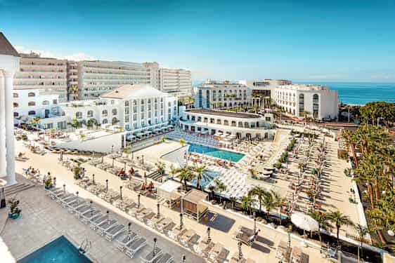 Utsikt över ett hotellområde med pooler, solstolar och vit arkitektur nära havet. (AI-genererad text)