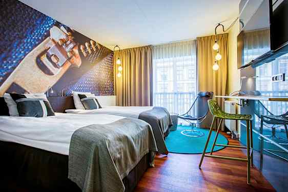 Ett modernt designat hotellrum med två enkelsängar, en illustrerad väggdekoration, gardiner och en fönsterutsikt. Rummet har också en grön stol, en svart stol och en väggmonterad TV. (AI-genererad text)