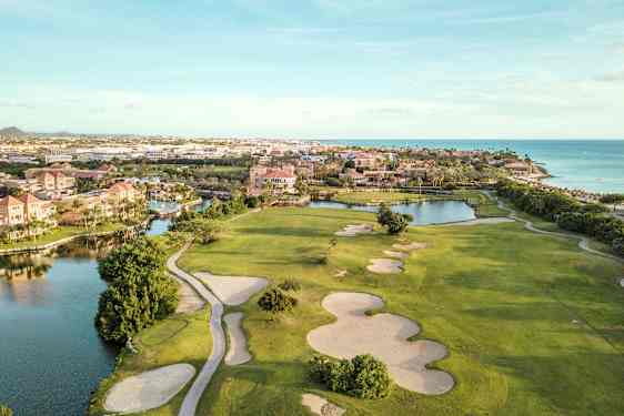 Golfbana med sandbunkrar och gröna gräsytor i närheten av vattendrag och strand. Bebyggelse i bakgrunden. (AI-genererad text)