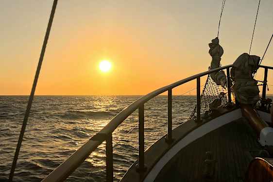 Solnedgång över havet med del av ett segelfartygs däck i förgrunden. (AI-genererad text)