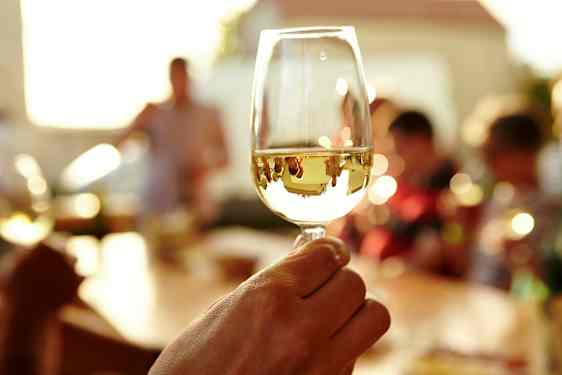 En person håller i ett glas vitt vin med en solbelyst bakgrund med människor. (AI-genererad text)