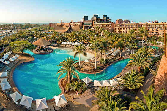 En stor utomhuspool omgiven av parasoller, solstolar och grönska vid ett semesterhotell. (AI-genererad text)