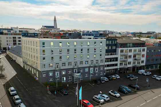 Flera moderna byggnader omger ett öppet parkeringsområde, med bilar parkerade på marken och en kyrka synlig i bakgrunden. (AI-genererad text)