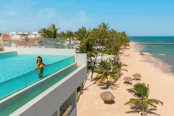 En pool med glasräcke på en byggnad med utsikt över en strand med palmer och azurblått hav. (AI-genererad text)