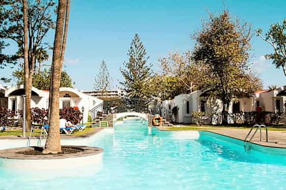 En pool med klart vatten och en liten bro, omgiven av grönska och semesterbostäder. (AI-genererad text)