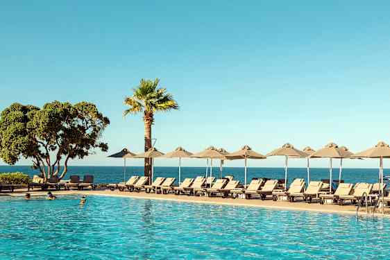 En pool i förgrunden med solstolar och parasoller vid sidan. Bakom syns havet och en klar blå himmel. (AI-genererad text)