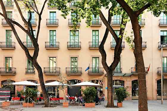Ett hotell med flera våningar och balkonger, en trottoar med parasoller och sittplatser bland träd. (AI-genererad text)