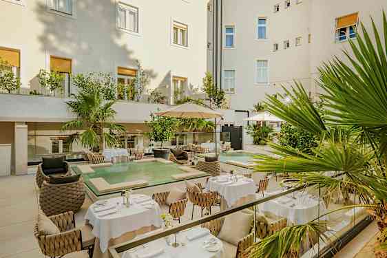 En mysig innergård med bord och stolar i en elegant miljö. Det finns också gröna växter och en liten pool. (AI-genererad text)