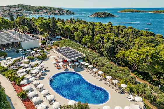 Vy över en utomhuspool omgiven av solstolar med parasoll, belägen nära kusten med utsikt över skog och hav. (AI-genererad text)