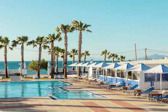 En pool med solstolar och parasoll under en klar himmel. Palmer och hav i bakgrunden. (AI-genererad text)