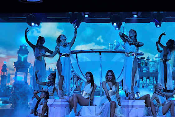 Dansare poserar på en upplyst scen med en dekor av palatsliknande byggnader mot en molnig himmel. (AI-genererad text)