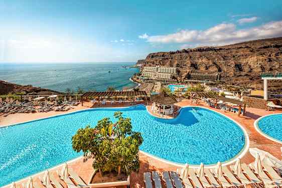 Hotellområde med pooler, solstolar och utsikt över havet samt omgivande klippor. (AI-genererad text)