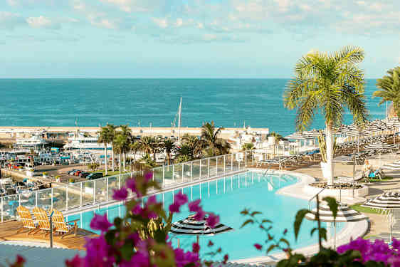 En pool med utsikt över ett hav och en hamn. I förgrunden finns växter och utemöbler. (AI-genererad text)