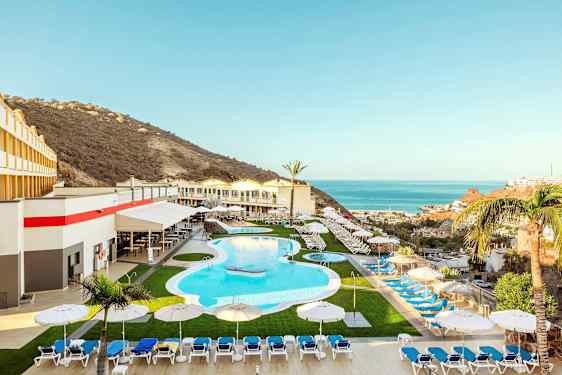 Ett semesterhotell med en pool, solstolar och parasoller, omgiven av en bergslandskap och utsikt över havet. (AI-genererad text)