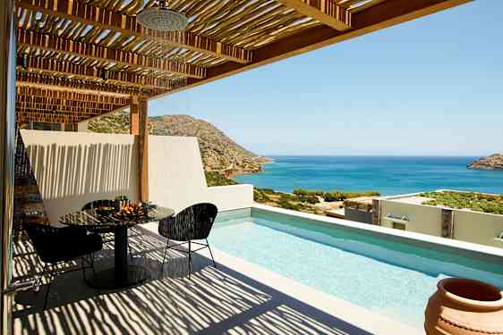 Utsikt över en terrass med pool, bord och stolar samt havet och bergen i bakgrunden. (AI-genererad text)