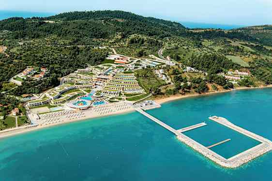 Flygbild över ett semesterområde med hotell, pooler och strand med solstolar, omgivet av grönska och hav. (AI-genererad text)