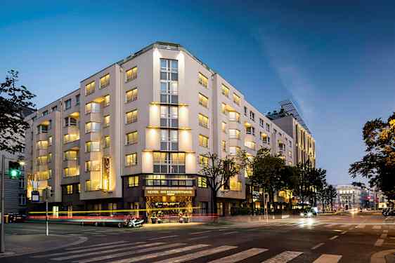 En byggnad som är upplyst på kvällen, med flera våningar och fönster, belägen vid en korsning. (AI-genererad text)