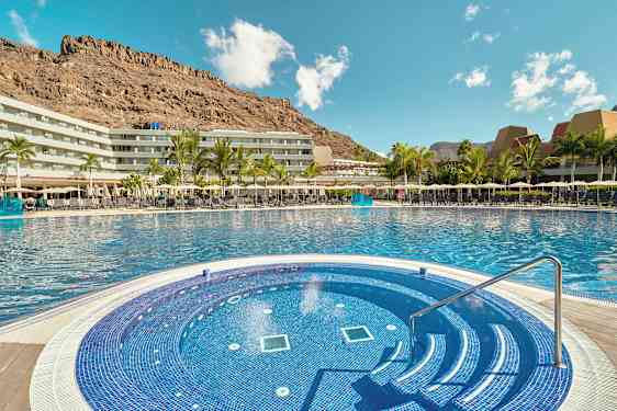 Hotellets poolområde med en mindre rund pool i förgrunden och omgivande byggnader samt berg i bakgrunden. (AI-genererad text)