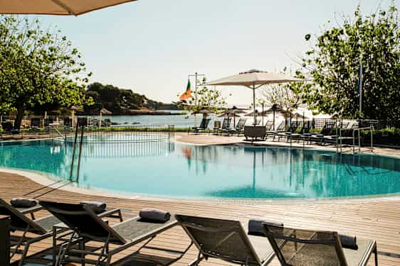 Pool med solsängar, träd och hav i bakgrunden. (AI-genererad text)