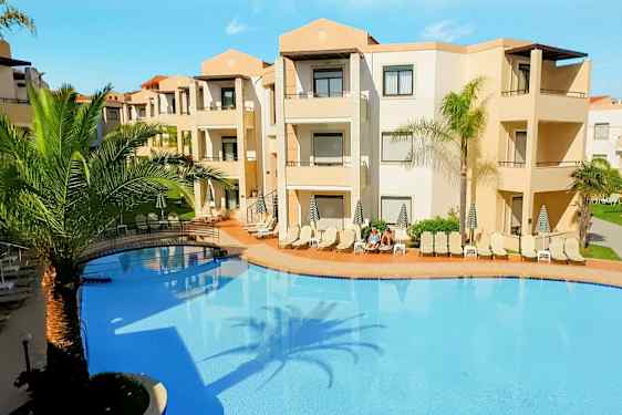 En utomhuspool omgiven av solstolar och palmträd vid en beige hotellbyggnad. (AI-genererad text)