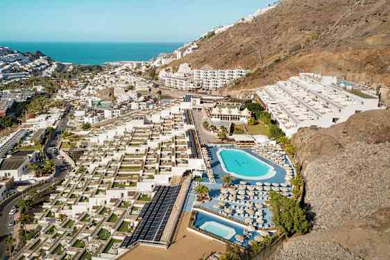 Flygfoto av en hotellanläggning med pooler, solstolar och terrasser, belägen vid en sluttning nära kusten. (AI-genererad text)