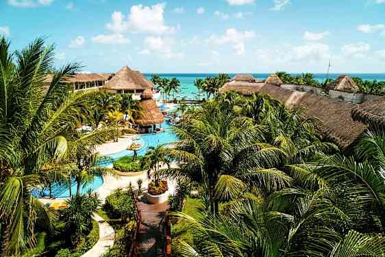 En stor resort med pooler, palmer och byggnader med halmtak, belägen nära havet under en blå himmel. (AI-genererad text)