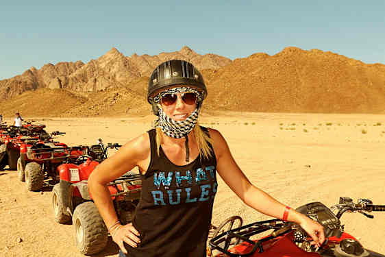 Kvinna står vid en röd fyrhjuling på en sandig ökenmark med berg i bakgrunden. (AI-genererad text)