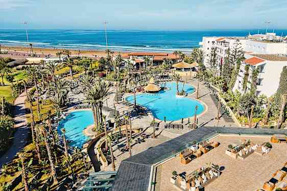 Hotellområde med pooler och omgivande trädgårdar, beläget nära en sandstrand och havet. (AI-genererad text)