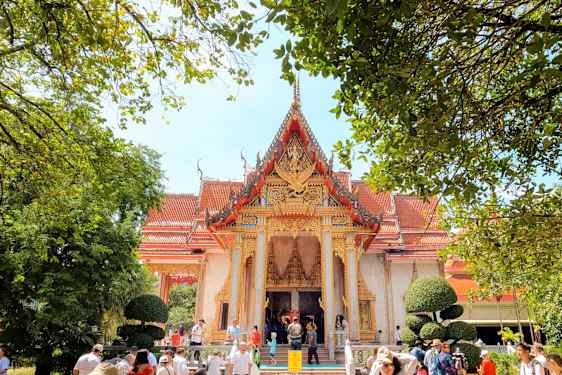 En traditionell byggnad med en dekorativ röd och guld takfront omgiven av gröna träd och människor. (AI-genererad text)