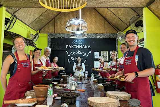 En grupp personer i förkläden står framför en skylt med texten 'Pakinnaka Cooking School' i en matlagningsstudio. (AI-genererad text)