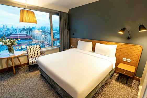 Ett hotellrum med en stor säng, fönster och utsikt över en stad. (AI-genererad text)