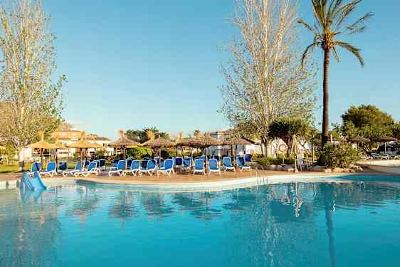 En pool vid ett hotell omgiven av solstolar och parasoller, omgärdad av träd och grönska. (AI-genererad text)