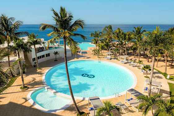 Ett hotellområde med en stor rund pool omgiven av palmer, solsängar och cabanas. Utsikt över havet i bakgrunden. (AI-genererad text)