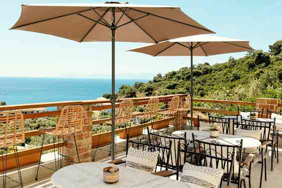 Terrass med bord, stolar och parasoller med utsikt över havet och grönskande omgivningar. (AI-genererad text)