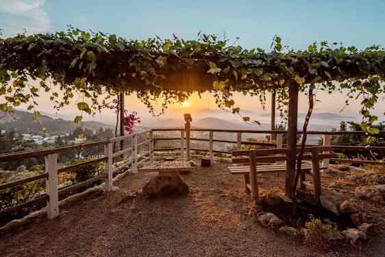 En utkiksplats med en träpergola täckt av löv samt bänkar och staket runtom. Solnedgång över ett bergigt landskap i bakgrunden. (AI-genererad text)