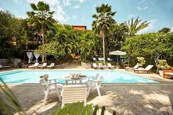 En utomhuspool omgiven av grönska och solstolar. Ett bord med mat och dryck står vid poolkanten. (AI-genererad text)