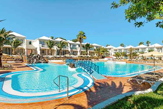 Ett hotell med vitmålade byggnader och en stor blå pool framför, omgiven av palmer och gröna områden. (AI-genererad text)