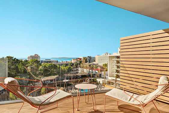 Vy över ett stadsmiljö med hotell och grönområden från en balkong med sittmöbler. (AI-genererad text)