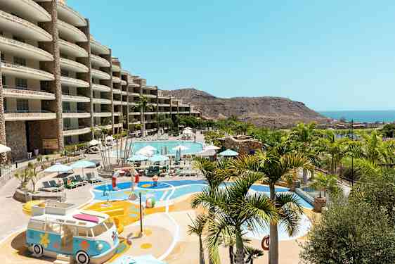 Utomhusområde med en stor pool, omgiven av solstolar och parasoller, samt lekplats med utsikt över havet och berglandskap. (AI-genererad text)