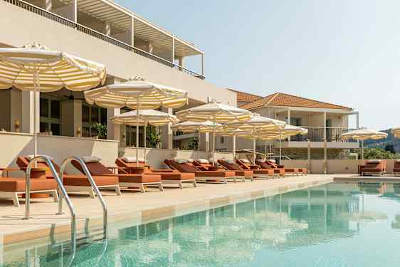 En pool omgiven av solstolar och parasoller vid en byggnad. (AI-genererad text)