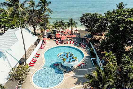 Flygbild över en pool vid en semesteranläggning med solstolar och parasoller, omgiven av tropiska träd och i närheten av havet. (AI-genererad text)